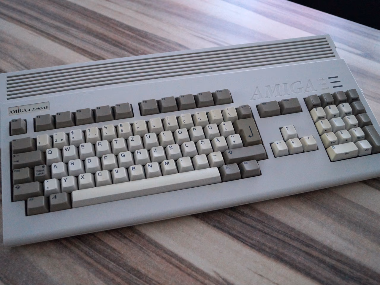 A gray Commodore Amiga 1200 on a wooden table, a wedge-shaped keyboard case with a numeric pad, the Amiga name molded into the top right and an A 1200 HD badge at the left.