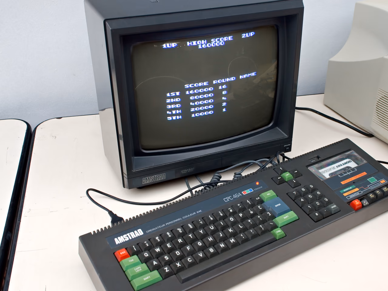 A dark gray Amstrad CPC 464 with a cassette deck built into the right of its keyboard, connected to an Amstrad color monitor showing a high-score table.