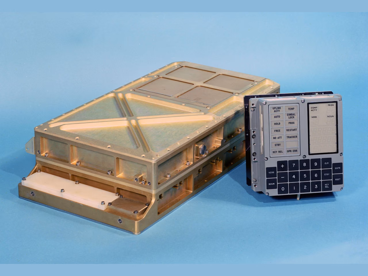 The Apollo Guidance Computer against a pale blue backdrop: a low rectangular metal case with a ribbed lid and bolted flanges along its sides, and standing beside it the display and keyboard unit, a panel of small caution lights beside blank display registers, with a square keypad below them.