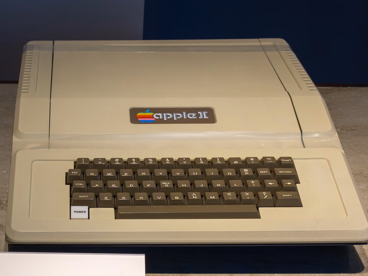 A beige Apple II seen from the front, its lid sloping down to an integrated keyboard with dark brown keys and a rainbow apple badge above them.