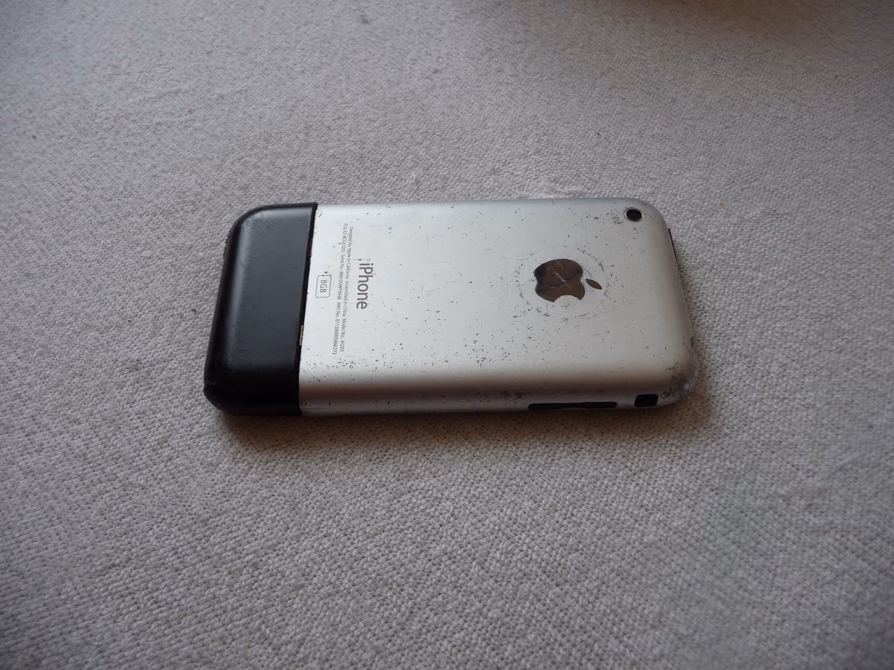 The back of a first-generation iPhone lying on a pale carpet: a brushed aluminum panel with a black plastic strip across the bottom, an Apple logo and the word iPhone engraved on it, and the metal scratched and scuffed with use.
