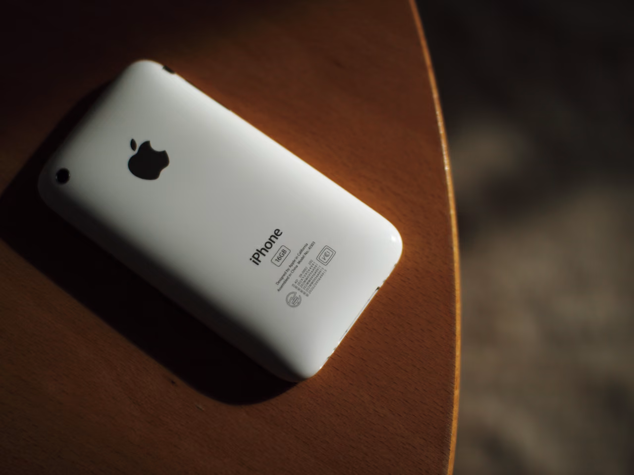 A white iPhone 3GS lying face down on a wooden table in a shaft of light: a curved glossy plastic back with an Apple logo, the word iPhone and the regulatory engraving below it.