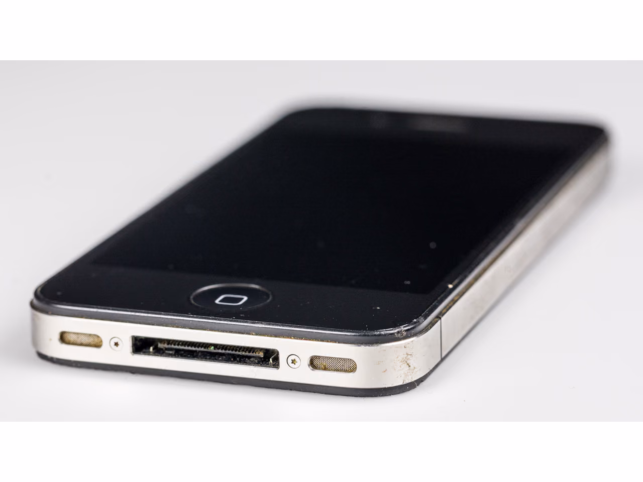 An iPhone 4 lying at an angle against white, seen from the front and one long side: a black glass face with a round home button, a stainless steel band around the edge carrying volume buttons, and the dock connector and speaker grilles along the bottom. The screen is off.
