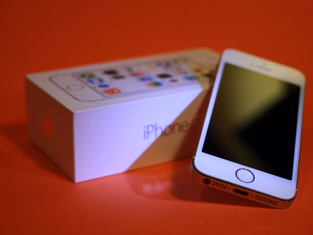 A gold iPhone 5s propped up against its white retail box on a red background: a black glass face with a round home button ringed in metal, and the charging port and speaker grilles along the bottom edge. The screen is off.