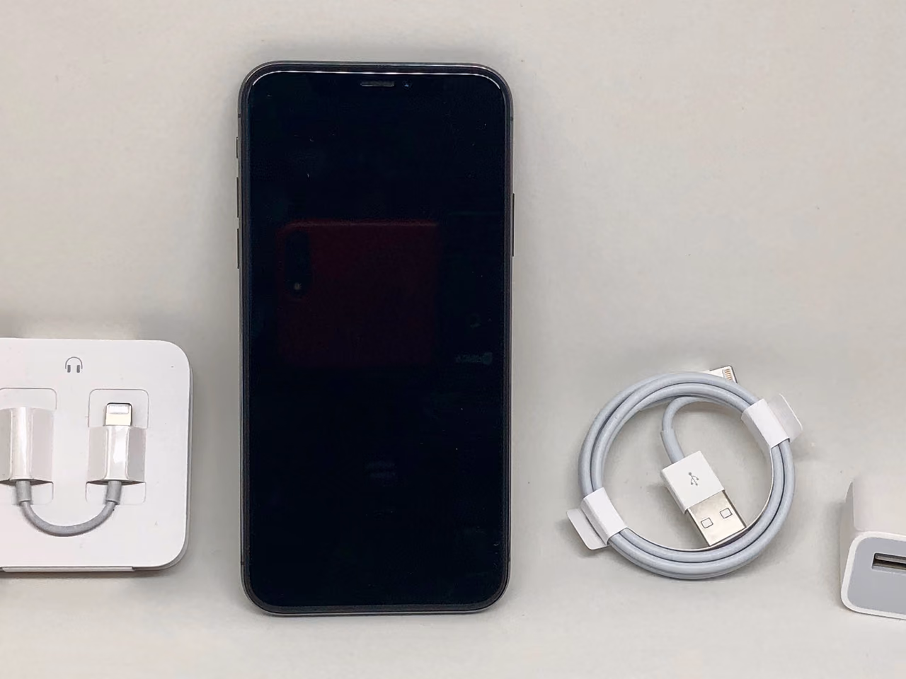 A space gray iPhone X seen face on against a pale surface: an all-glass front with the notch at the top and no home button, and the headphone adapter, charging cable and power adapter it shipped with laid out on either side. The screen is off.