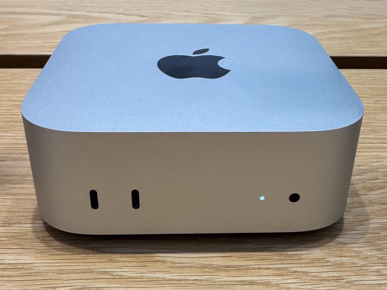 A small square aluminum Mac mini on a wooden table, an Apple logo on its top face and two USB-C ports with a status light on the front edge.