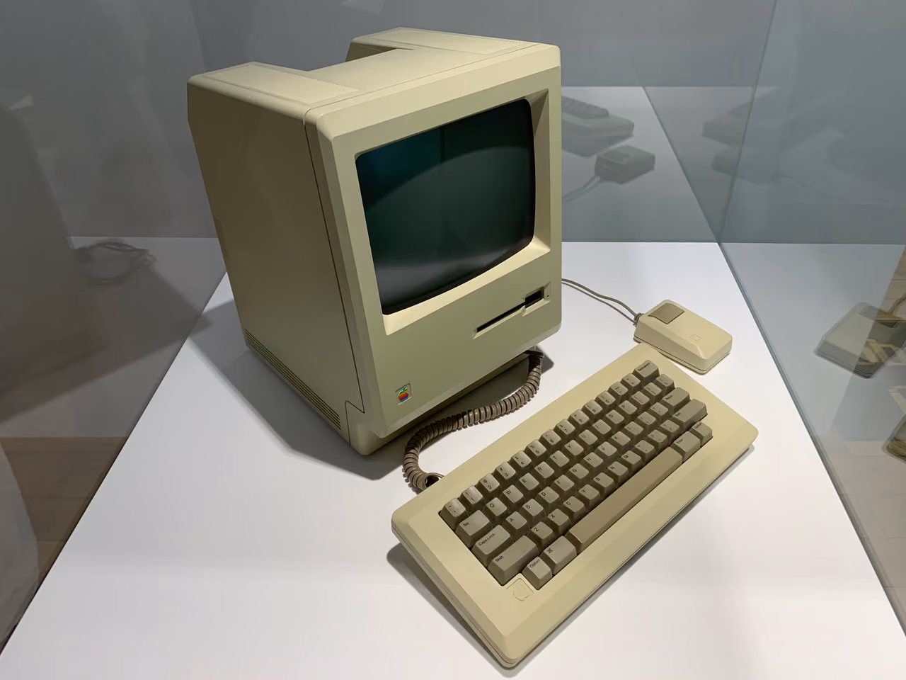 A beige Macintosh 128K standing on a white plinth, its small screen above a diskette slot, with the coiled-cable keyboard in front of it and a single-button mouse to the right.