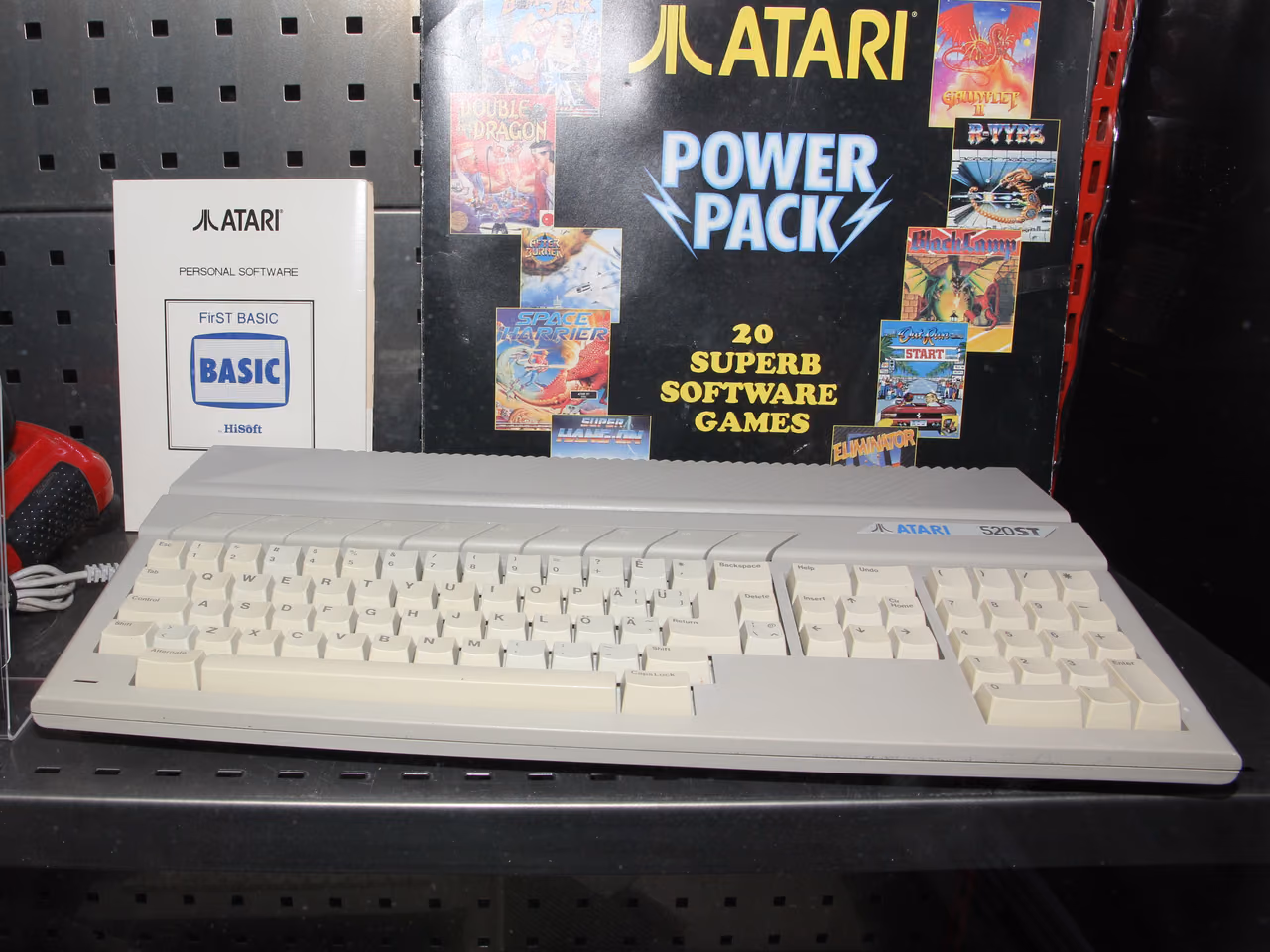 A cream Atari 520ST on a museum shelf: a wide keyboard unit with a numeric pad at the right and the Atari logo and model name printed above it, with a boxed games compilation standing behind.