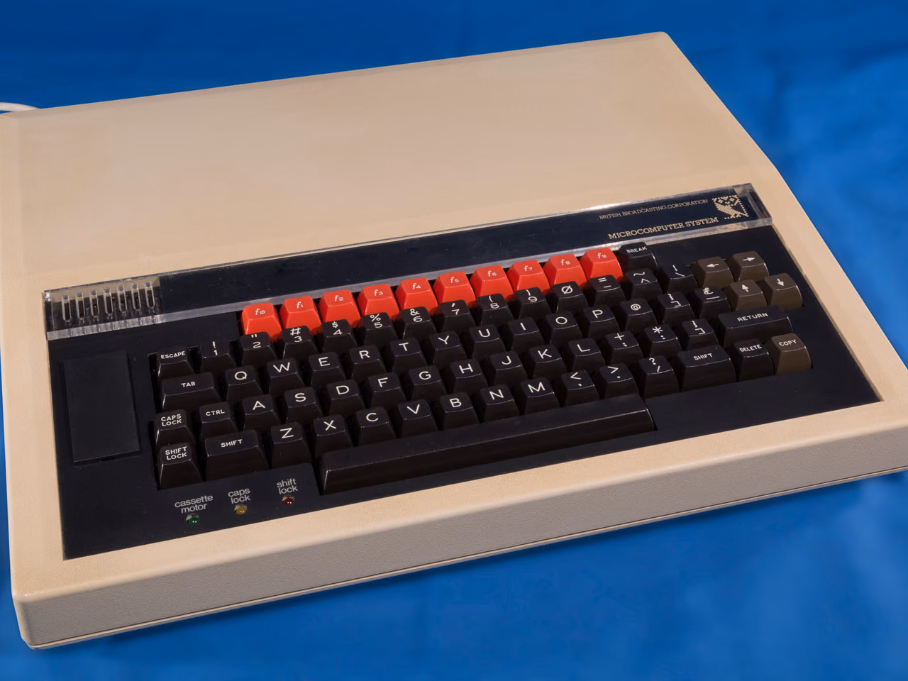 A cream BBC Micro on a blue cloth, its black keyboard crossed by a row of red function keys and the BBC owl badge at the top right of the case.