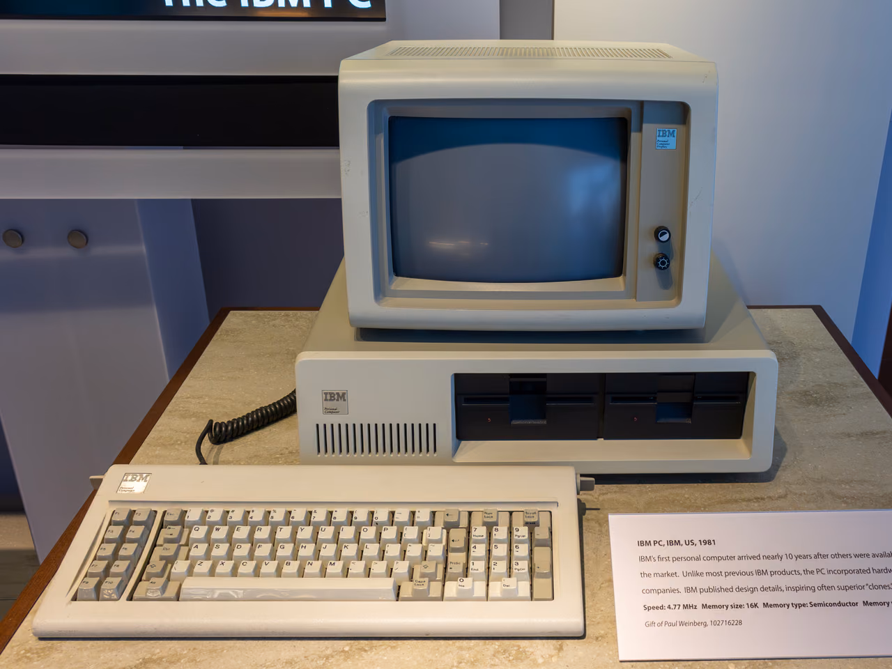 An IBM Personal Computer on a museum plinth: a wide beige system unit with two diskette drives, a monochrome monitor standing on top of it and the keyboard in front.