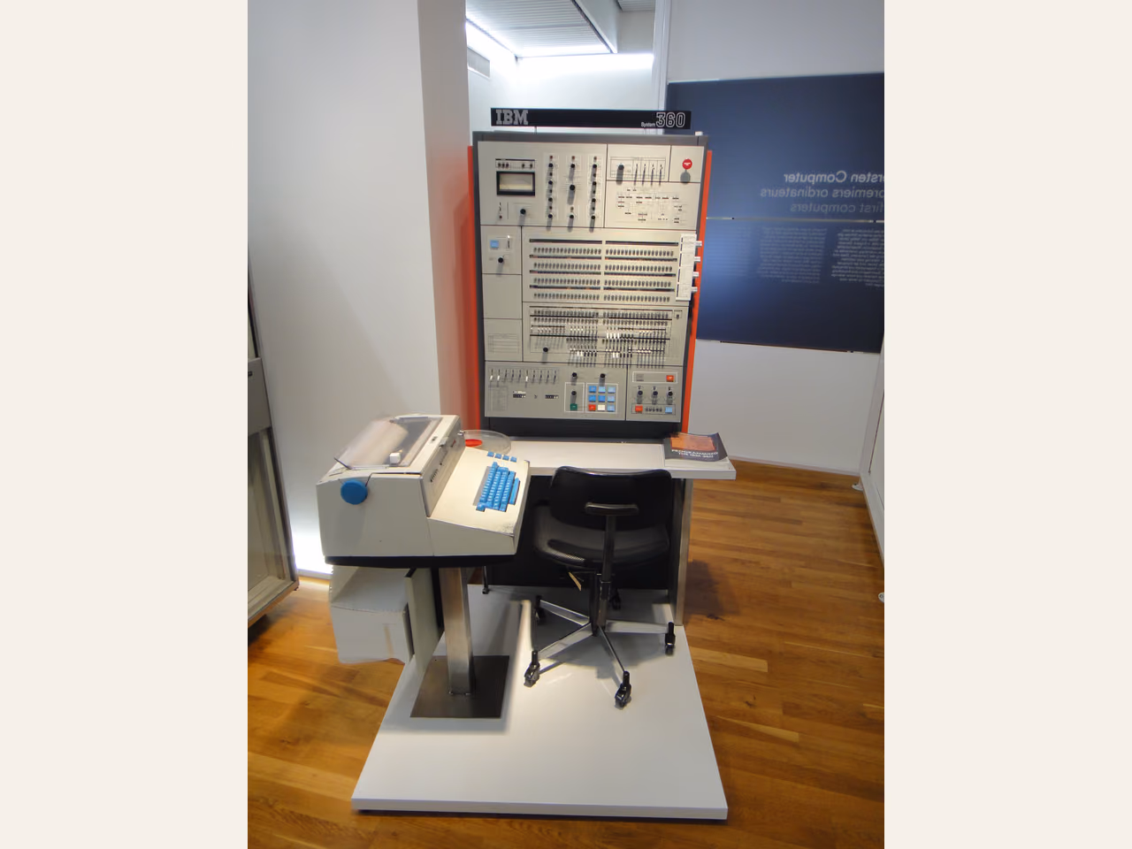 An IBM operator panel with rows of indicator lights and switches, above a desk containing a printer and keyboard.
