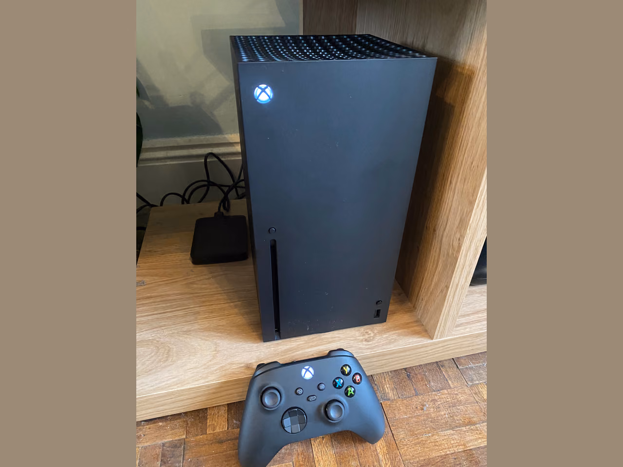 An Xbox Series X standing upright on a wooden shelf: a tall matte black box with a grille of ventilation holes across the top, a lit white Xbox logo near the top of the front face, a disc slot down its left edge, and the black controller lying on the floorboards in front of it.