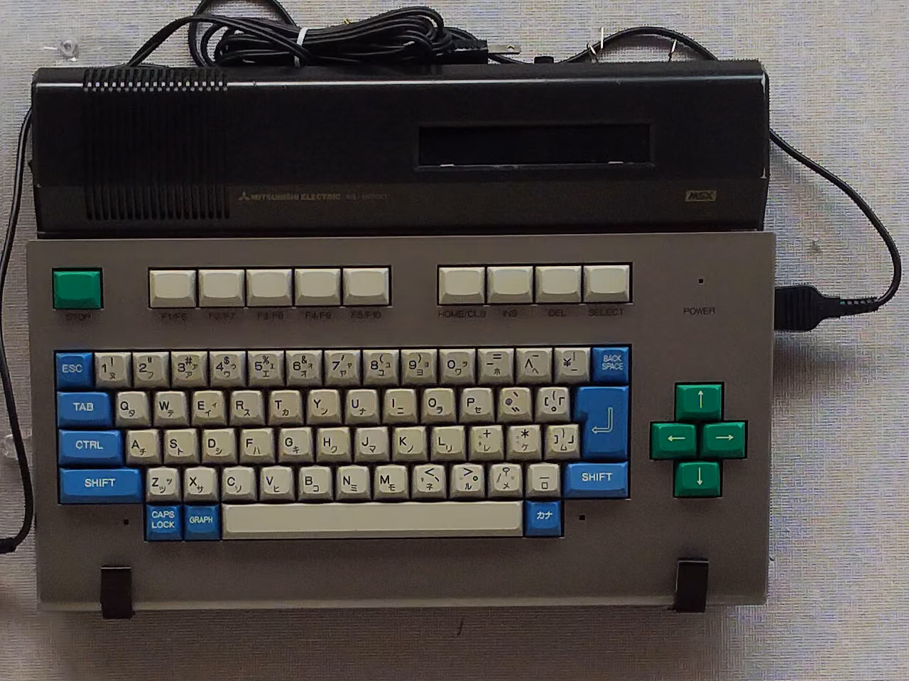 A Mitsubishi ML-8000 mounted on a display panel: a gray keyboard unit with blue and green keys, under a black upper section holding a cartridge slot.