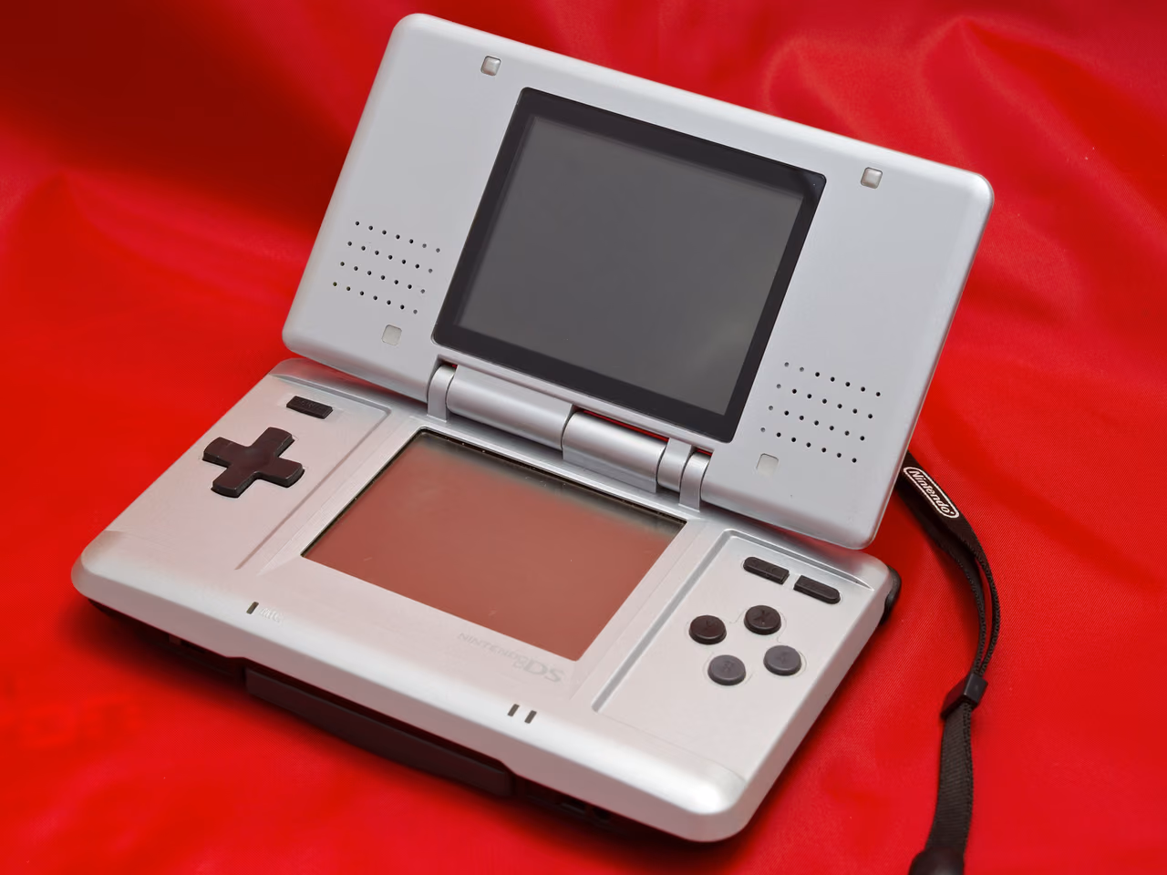 A first-generation Nintendo DS opened out on red cloth: a silver clamshell with one screen in the lid and a second in the base, a cross-shaped pad at the left of the base, four buttons at the right, and a wrist strap looped away to the side. Both screens are off.