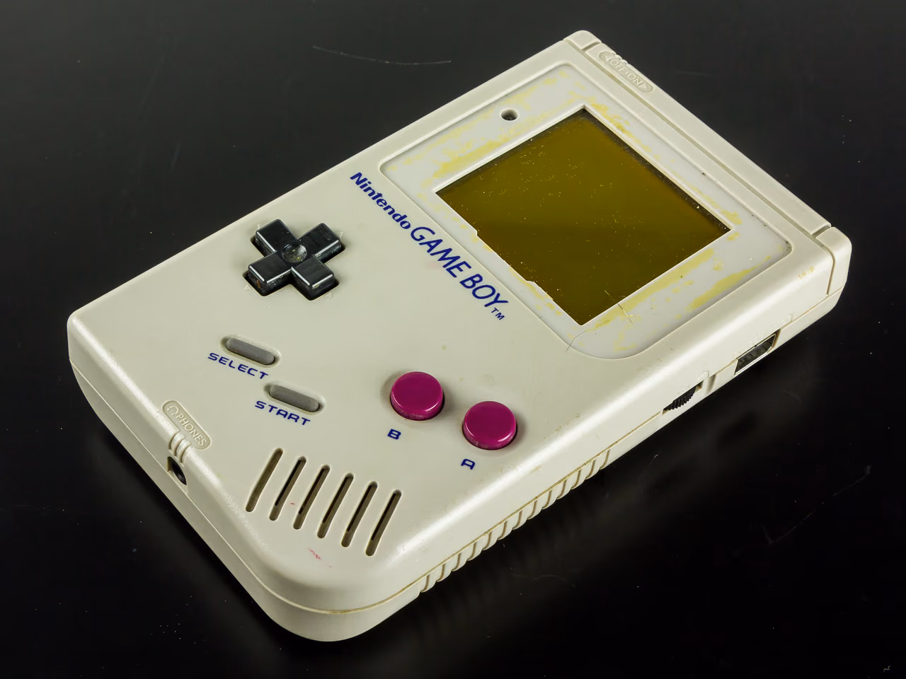 A Nintendo Game Boy on a black surface, seen from above at an angle: a gray handheld with a dark green screen window, a black cross-shaped pad, two magenta buttons marked B and A, and SELECT and START below the pad. The plastic has yellowed.