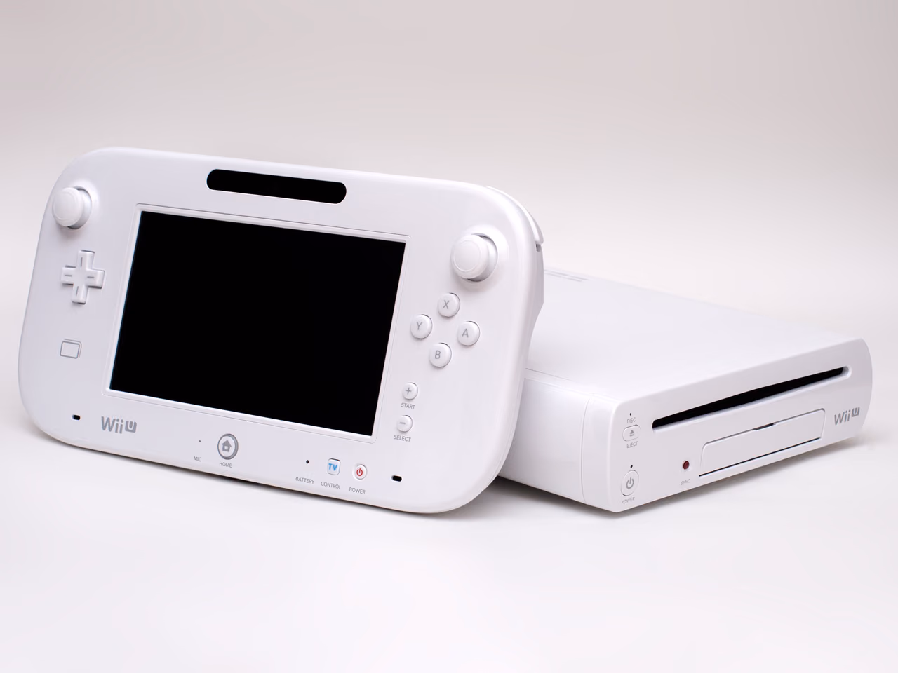 A white Wii U GamePad, its screen switched off, standing in front of the white Wii U console, which lies flat behind it with the disc slot facing forward.