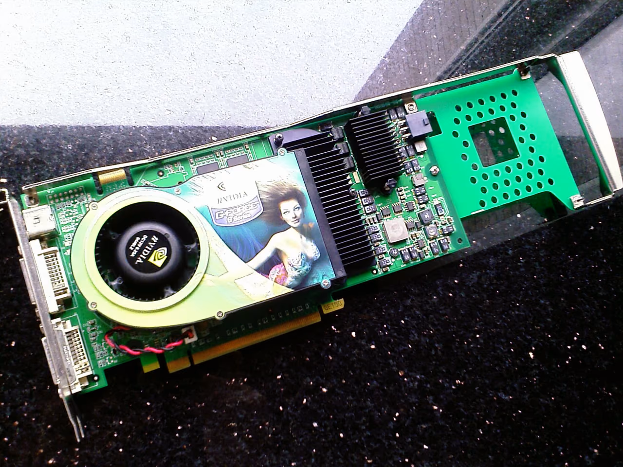 A GeForce 6800 Ultra lying on dark ground: a long green board carrying a green cooler with printed GeForce 6 artwork over its fan, a black finned heatsink beside it, and two DVI sockets on the bracket.