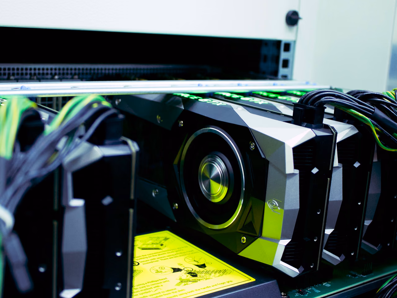 A row of NVIDIA GeForce GTX 1080 Founders Edition cards mounted side by side in a rack, the nearest one in focus with its angular silver shroud and centrifugal fan lit green, power cables looping away from their connectors.