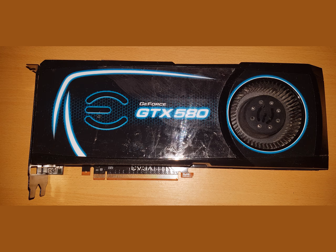 An EVGA GeForce GTX 580 seen from above on a wooden table: a black shroud printed with the EVGA logo and the model name, a radial fan at one end, and the PCI Express edge connector along the bottom.