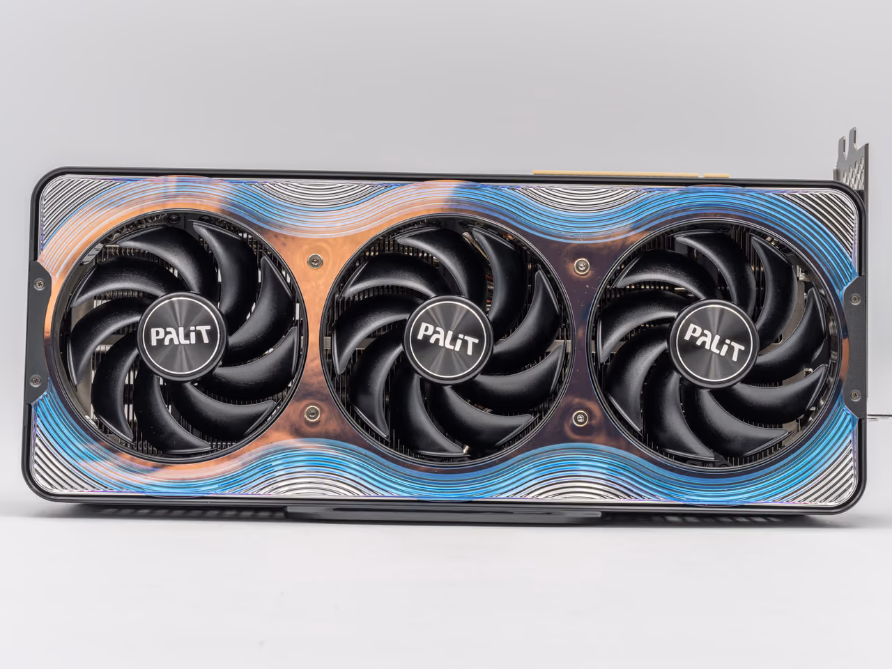 A Palit GeForce RTX 5090 GameRock seen face on against pale gray: three black fans set into a shroud patterned with wavy blue, copper and white lines, with the mounting bracket standing out at one end.