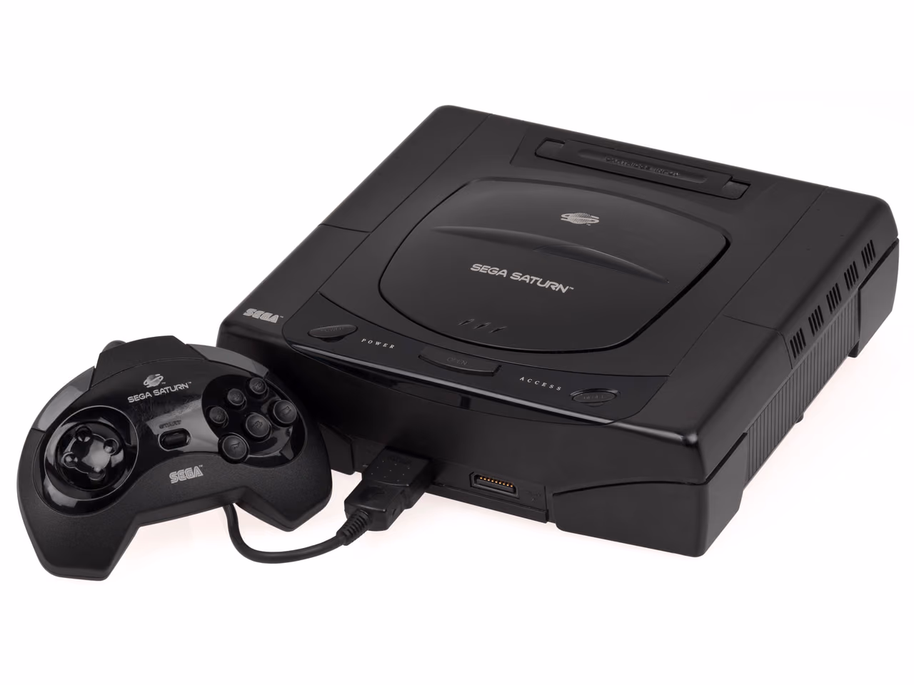 A black Sega Saturn photographed against white, a circular disc lid recessed into the top of the case, with the first-model control pad lying in front of it.