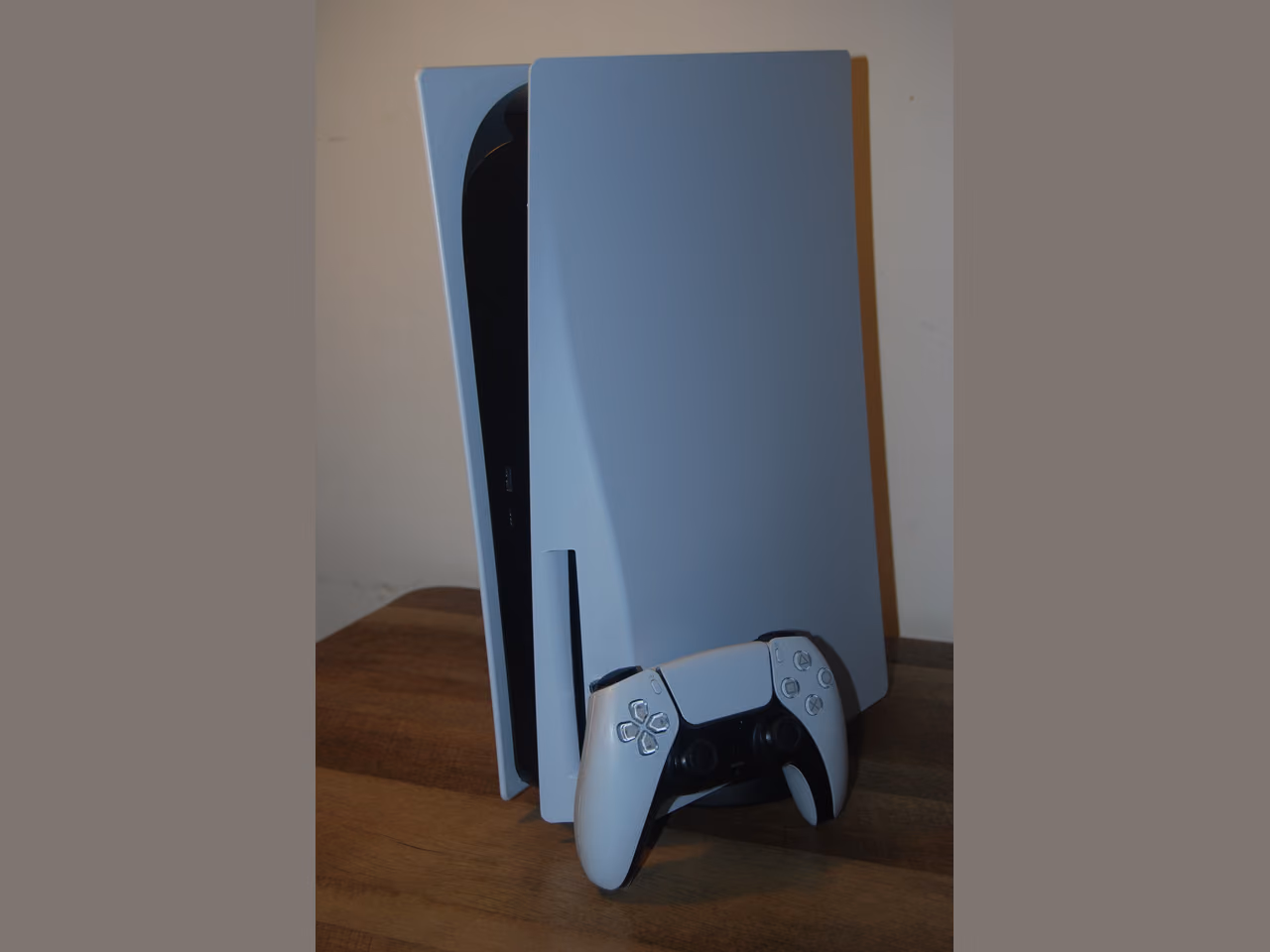 A PlayStation 5 standing upright on its stand on a wooden table: two curved white side panels around a narrower black center, a disc slot low on the front, and the white DualSense controller resting against the base.