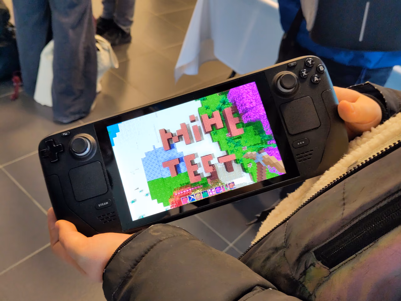 A Steam Deck held in both hands: a wide black handheld with a screen in the middle, thumbsticks, a cross-shaped pad and trackpads at the left, and four buttons and a second trackpad at the right. The screen shows a blocky landscape with the word Minetest across it.